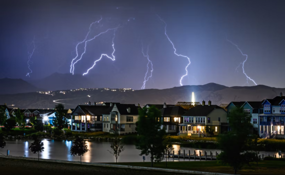 A row of houses with a lake in front with lightning flashes in the background