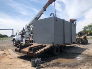 A large gray storm shelter or safe room is being loaded or unloaded from a flatbed trailer using a crane, with workers assisting on-site.