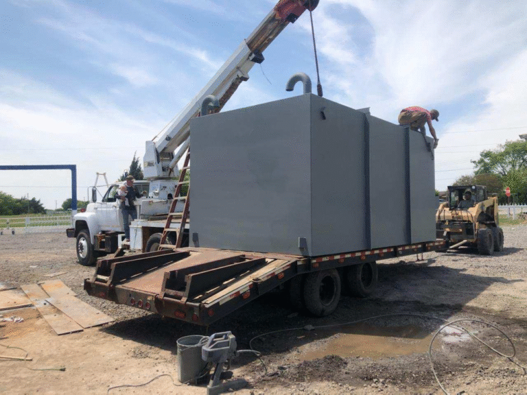 A large gray storm shelter or safe room is being loaded or unloaded from a flatbed trailer using a crane, with workers assisting on-site.