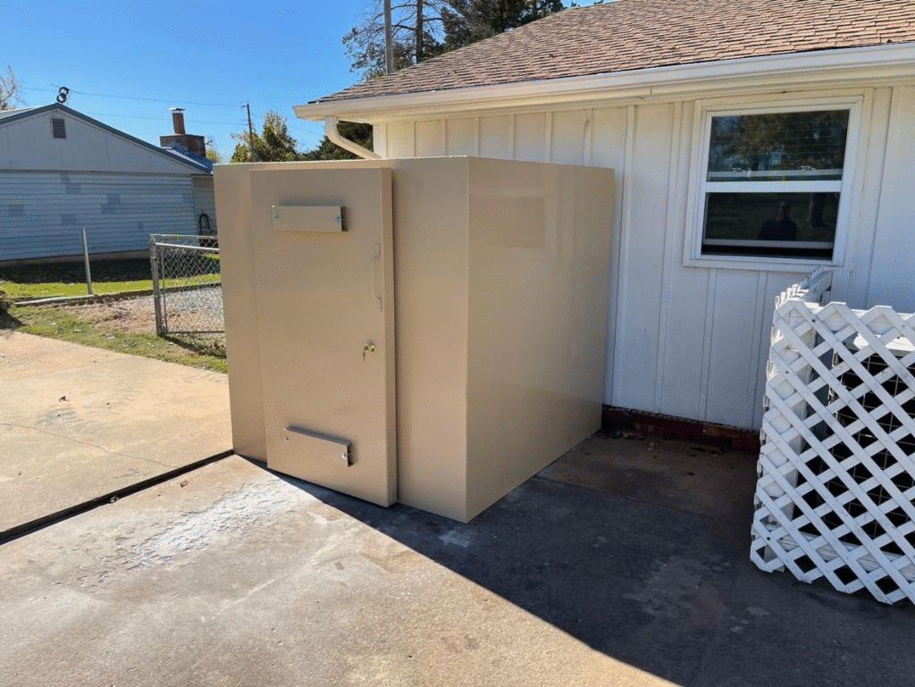 The image shows a beige outdoor storm shelter or safe room installed against the exterior wall of a house on a concrete surface.