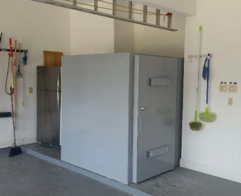 The image shows a large, industrial-style safe installed in a garage with cleaning tools hanging nearby.