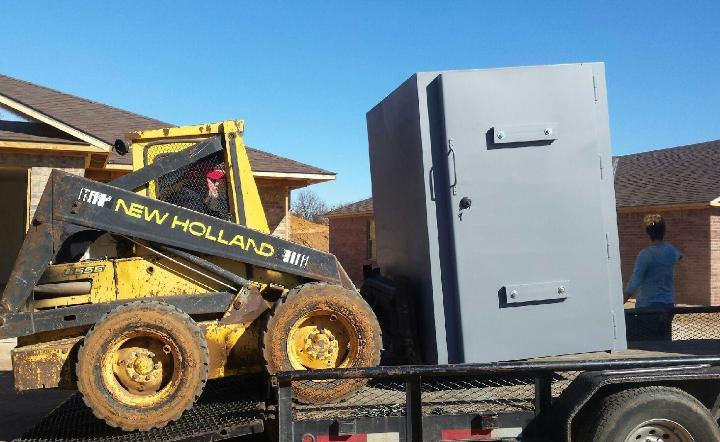 Heavy-duty steel storm shelter being transported on a trailer with a skid steer at a residential property