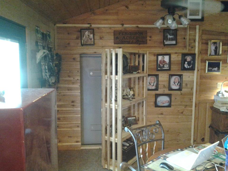 A gray steel safe room door built into a wood-paneled interior wall, surrounded by shelving and family photos.