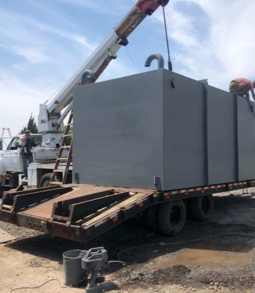 A large gray steel storm shelter being lowered by a crane onto a flatbed trailer at an outdoor site, with two workers present.
